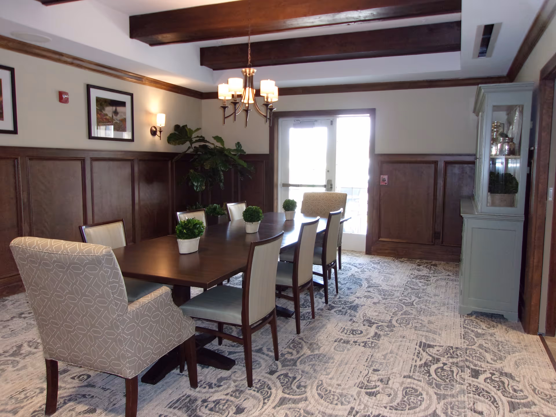 A dining room with a long wooden table surrounded by eight chairs, two of which have patterned upholstery. The room features wood paneling on the walls, a patterned carpet, a chandelier hanging from a ceiling with exposed wooden beams, framed pictures on the walls, a large plant in the corner, and a glass-front cabinet on the right side. Double glass doors let in natural light from outside.