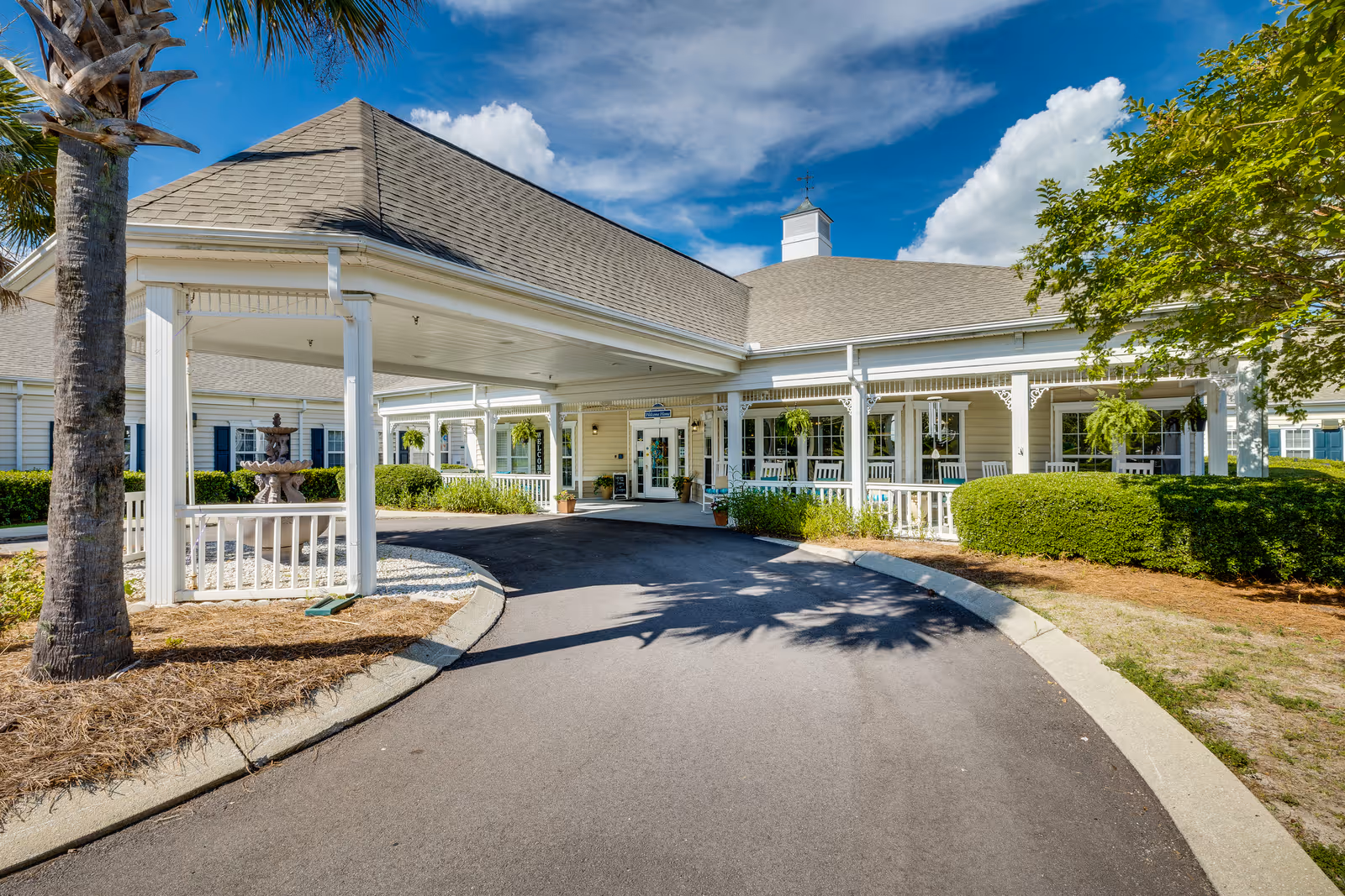 Front exterior view of The Legacy of Lexington facility showing a covered driveway entrance with white columns, a fountain on the left side, manicured bushes, and a clear blue sky with some clouds.