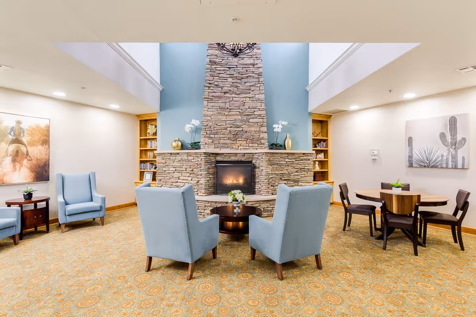 A cozy living room area with a tall stone fireplace in the center. Four light blue armchairs are arranged around a small round wooden table in front of the fireplace. To the right, there is a round wooden dining table with four dark chairs. The walls are decorated with framed artwork, and built-in wooden bookshelves flank the fireplace. The room has a patterned carpet and soft lighting from recessed ceiling lights.