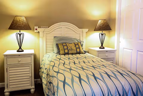 A cozy bedroom featuring a single bed with a patterned bedspread and decorative pillows. The bed has a white wooden headboard and is flanked by two white nightstands, each with a lamp emitting warm light. The walls are painted a soft beige color, and there is a white door visible on the right side.