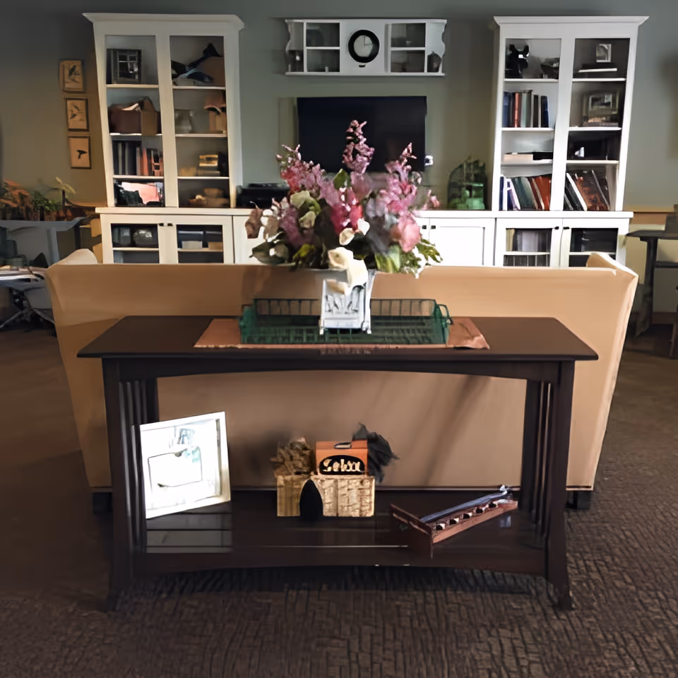 A cozy living room area with a beige sofa facing a dark wooden console table adorned with a floral arrangement. Behind the sofa, there are white built-in shelves filled with books and decorative items, and a wall-mounted TV in the center.