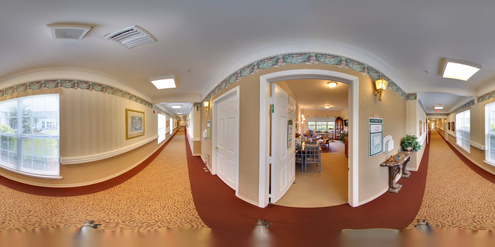 A wide hallway in a senior living facility with patterned carpet and wallpaper. The hallway has large windows on both sides letting in natural light. There is an open doorway leading into a cozy living room area furnished with chairs, tables, and lamps. The walls are decorated with framed artwork and a decorative border near the ceiling. A small table with plants and informational signage is visible near the open doorway.
