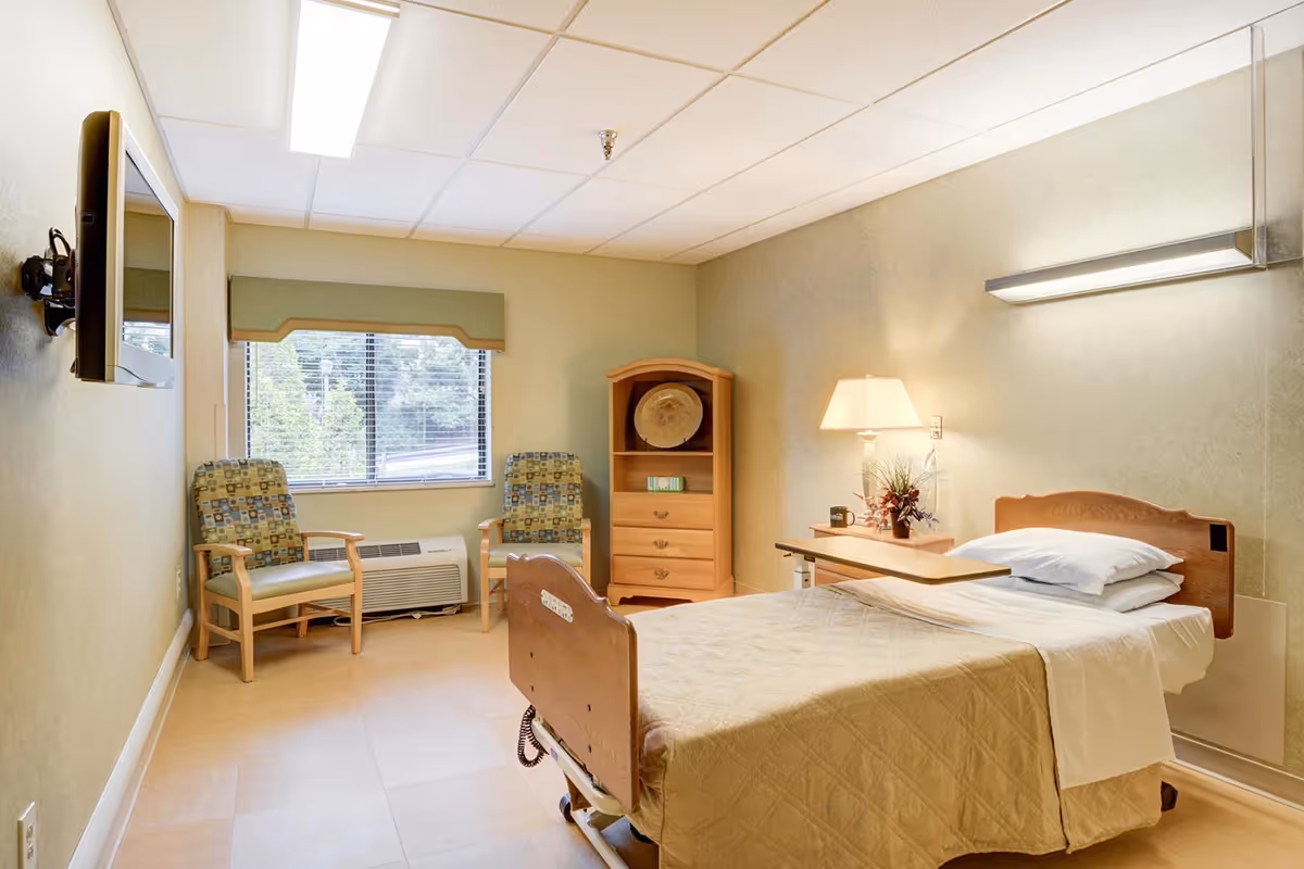 A patient room in a senior living facility with a single bed, two chairs by the window, a dresser, and a bedside lamp.