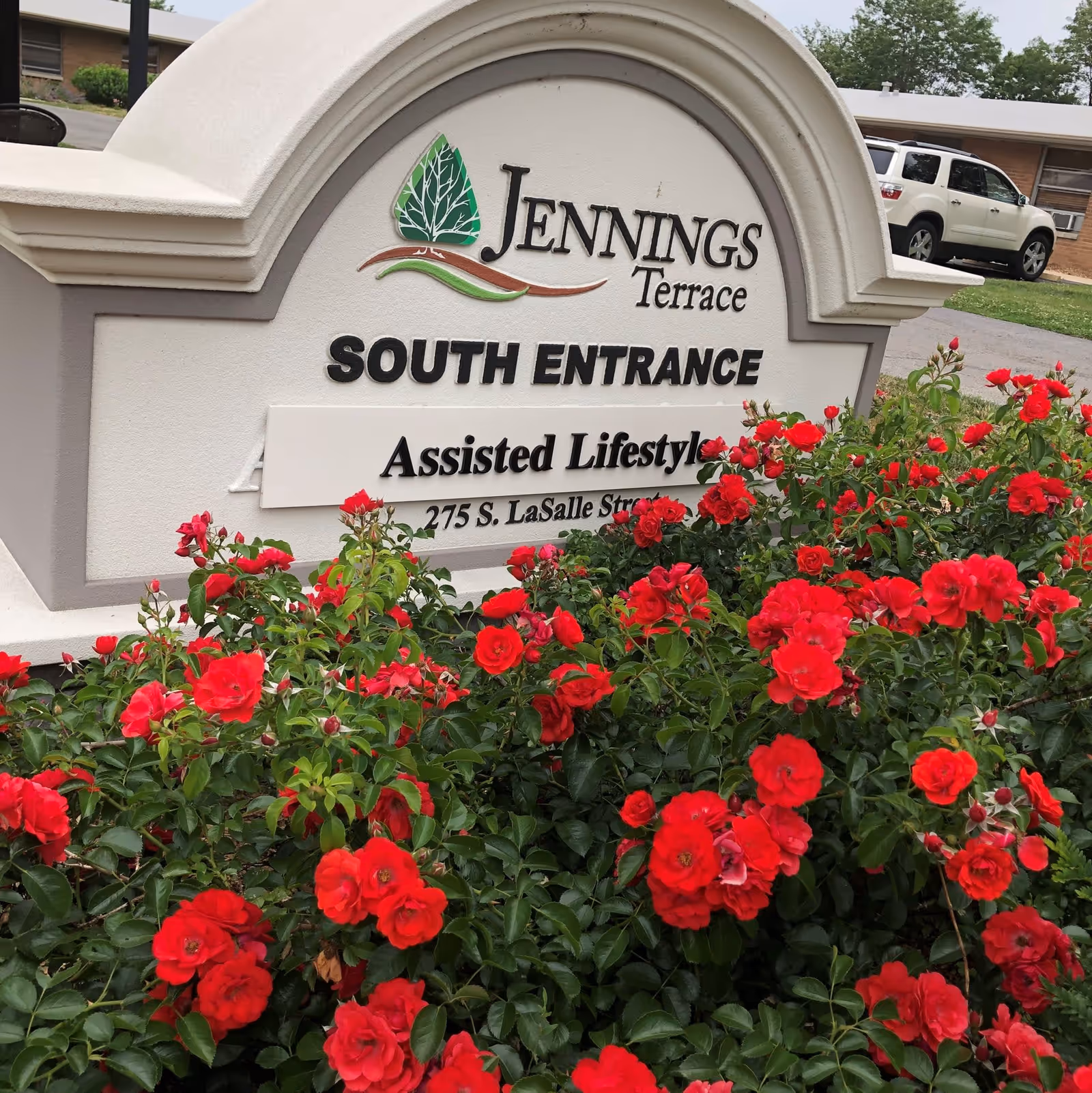 A stone sign for Jennings Terrace South Entrance with red flowers in the foreground and a white SUV parked in the background near a building.