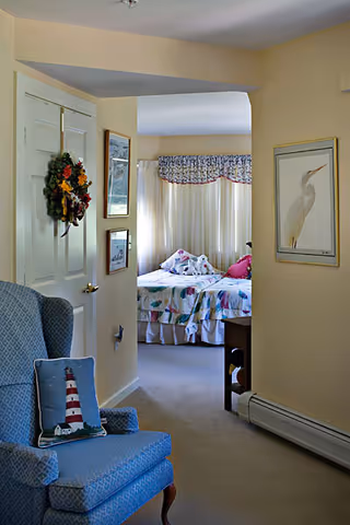 View of a cozy interior space showing a blue upholstered armchair with a lighthouse pillow in the foreground, a door decorated with a floral wreath, and a bedroom in the background with a bed covered in floral bedding and stuffed animals. The walls are light yellow, and there is framed artwork including a picture of a bird.