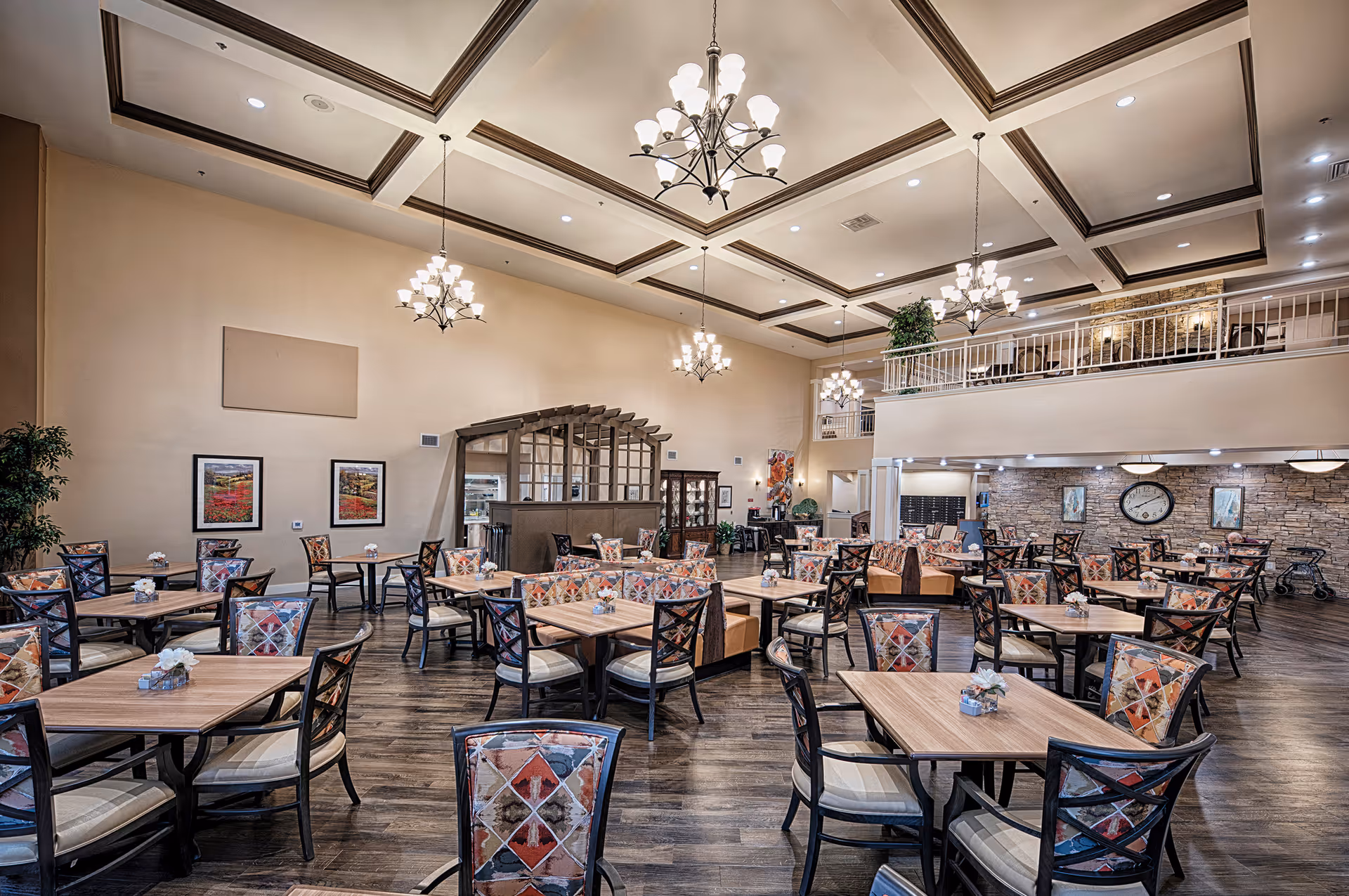 Spacious dining room with multiple wooden tables and chairs featuring colorful patterned upholstery. The room has high ceilings with decorative beams and multiple chandeliers. There is a stone accent wall with a large clock and framed artwork, and a second-floor balcony overlooking the area. The floor is dark wood, and the room is well-lit with recessed lighting.
