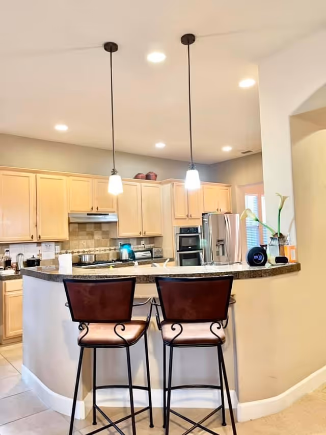 A modern kitchen with light wood cabinets, a granite countertop island with two high chairs, stainless steel appliances including a refrigerator and oven, pendant lights hanging from the ceiling, and a small digital clock and vase with flowers on the counter.