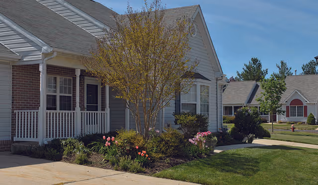 Exterior view of a residential-style senior living facility with a small porch, landscaped garden with flowers and shrubs, and a tree in front. Other similar buildings and a fire hydrant are visible in the background under a clear blue sky.