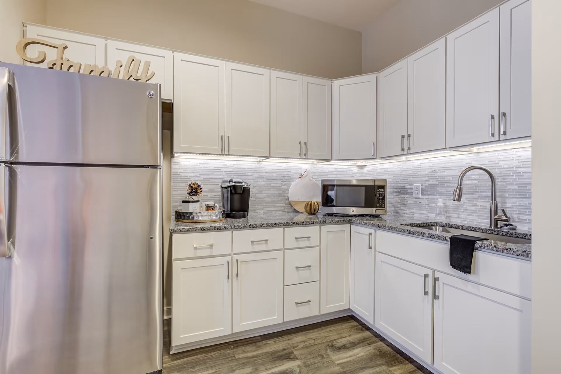 Modern kitchenette with stainless steel refrigerator, white cabinets, granite countertops, microwave, sink and under-cabinet lighting.