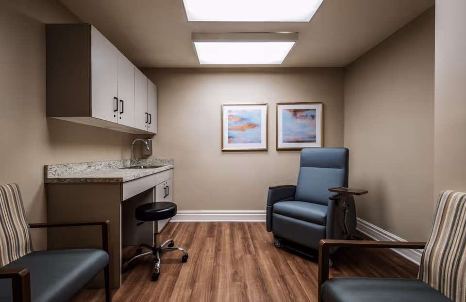 A small interior room with a reclining medical chair, visitor chairs, a sink and cabinets, and two framed paintings on the wall.