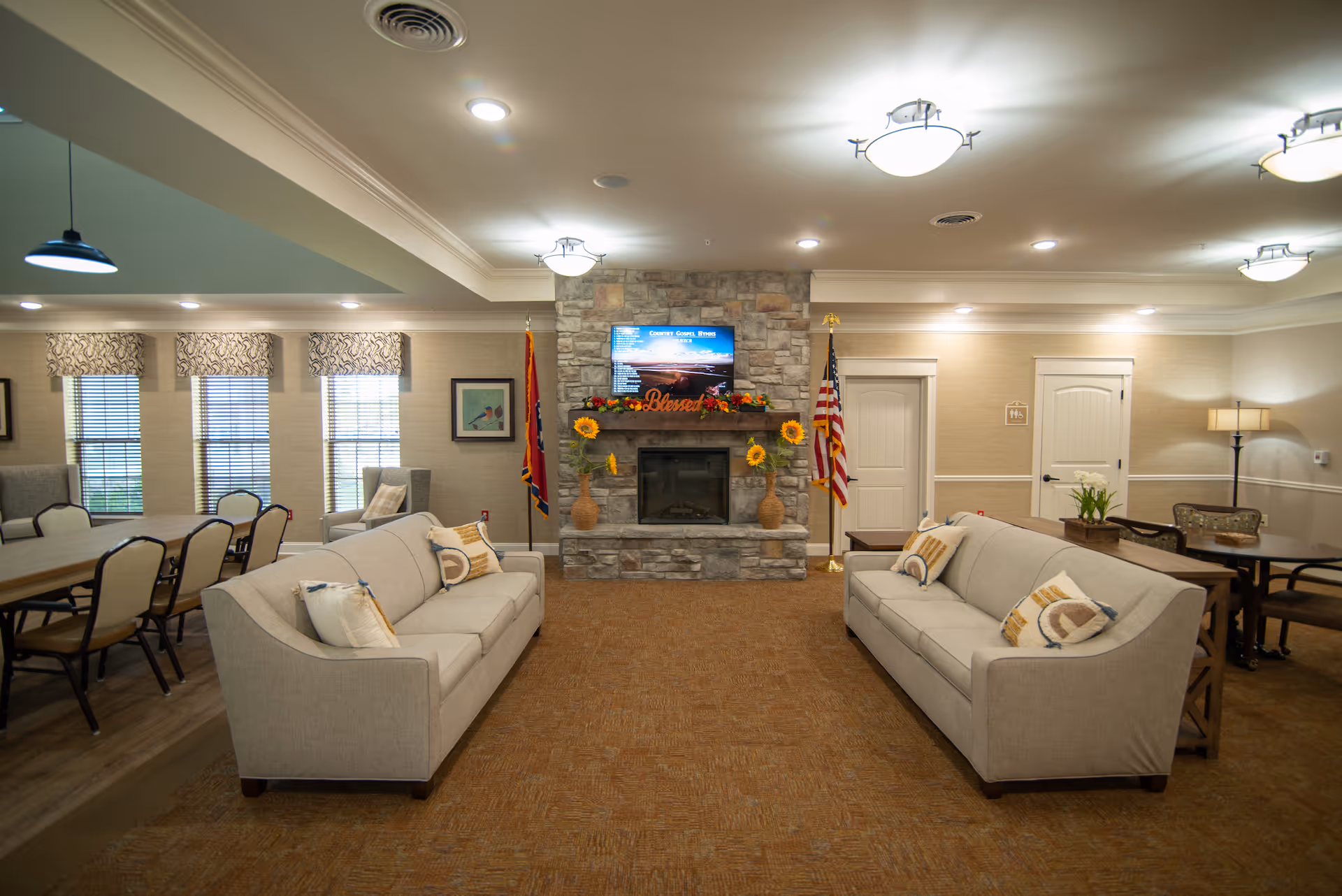 A cozy living room area with two beige sofas facing each other, each adorned with decorative pillows. In the center background is a stone fireplace decorated with sunflowers and a 'Blessed' sign, with a flat-screen TV mounted above it. To the left, there is a dining area with a long table and chairs, and windows with patterned valances. The room is warmly lit with ceiling lights and a floor lamp in the corner. The American flag and another flag stand on either side of the fireplace.