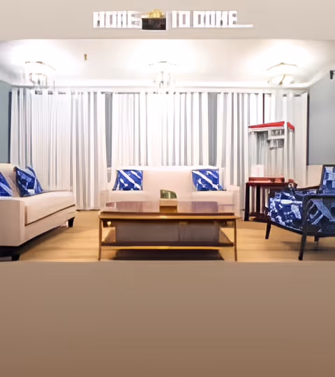 Well-lit communal living room with sofas, patterned pillows, armchairs, a central coffee table and vertical blinds along the back wall.