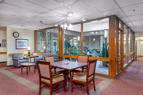 A common area in a senior living facility featuring a square wooden table with four upholstered chairs. Behind the table is a glass-enclosed indoor garden with various green plants. To the left, there is a small sitting area with a patterned armchair, a round wooden side table, a lamp, and framed artwork on the wall. The floor is carpeted in a red pattern, and the ceiling has recessed lighting.