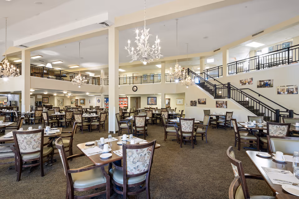 Spacious dining room in a senior living community with multiple tables and chairs arranged neatly. The room features high ceilings, chandeliers, and a staircase leading to an upper level with railings. Tables are set with white cups, plates, and silverware. The walls are decorated with framed pictures and the carpet has a subtle pattern.