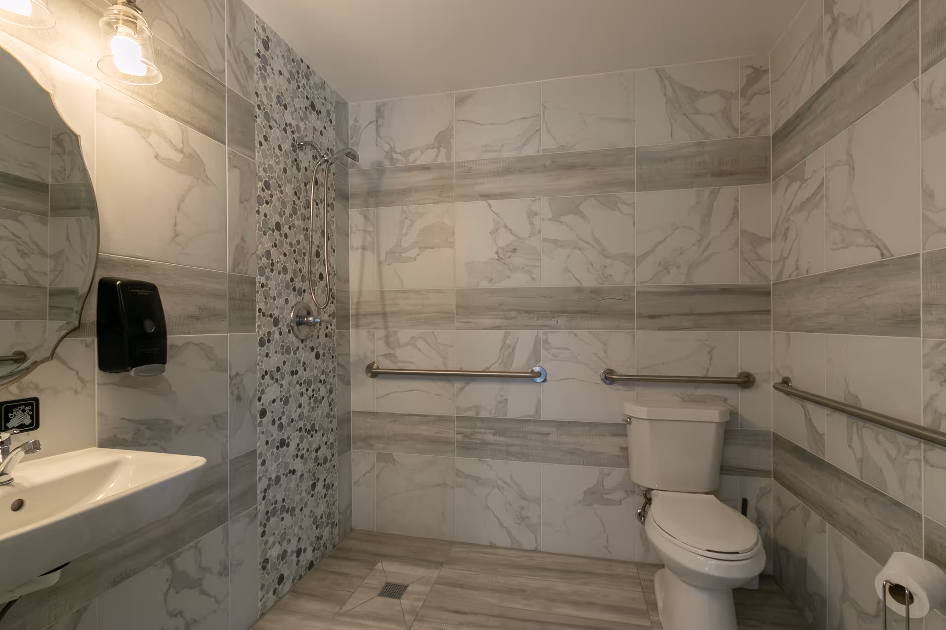 A senior-friendly bathroom with marble-patterned tiles on the walls and floor, featuring a wall-mounted sink with a soap dispenser, a toilet with grab bars on both sides, and a walk-in shower area with a handheld showerhead and pebble-style accent tiles.