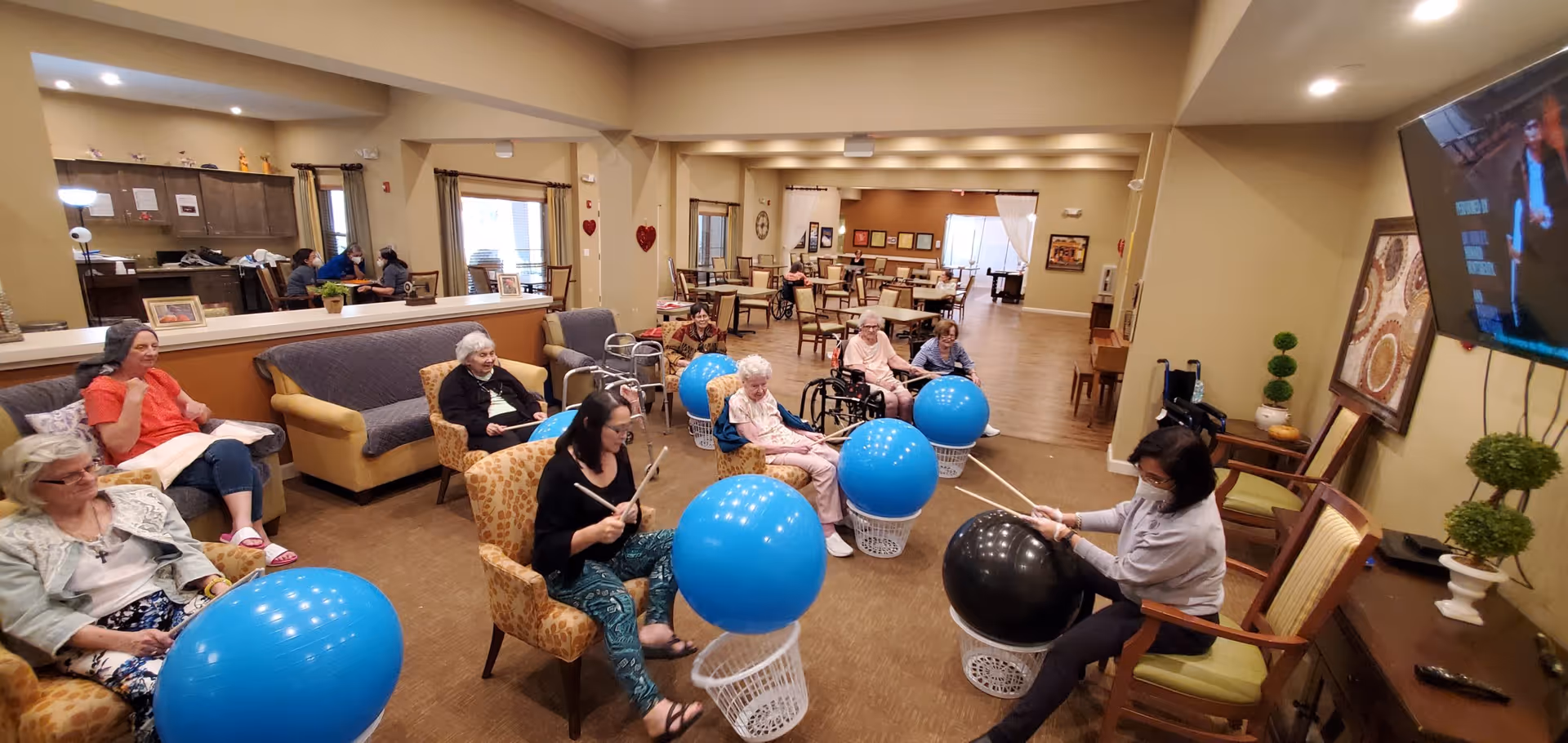 A group of elderly individuals seated in a common room participating in a drumming exercise using large blue and black exercise balls placed on white baskets. Two facilitators are guiding the activity. The room has beige walls, comfortable chairs, a large TV mounted on the wall, and a dining area visible in the background.