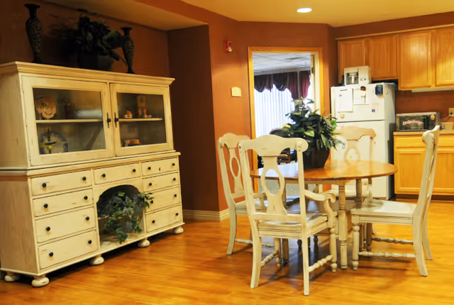 A cozy dining area with a round wooden table surrounded by four white wooden chairs. Behind the table is a white wooden cabinet with glass doors displaying decorative items and plants. The room has warm-colored walls and wooden flooring. In the background, there is a kitchen area with wooden cabinets, a refrigerator, and a microwave.
