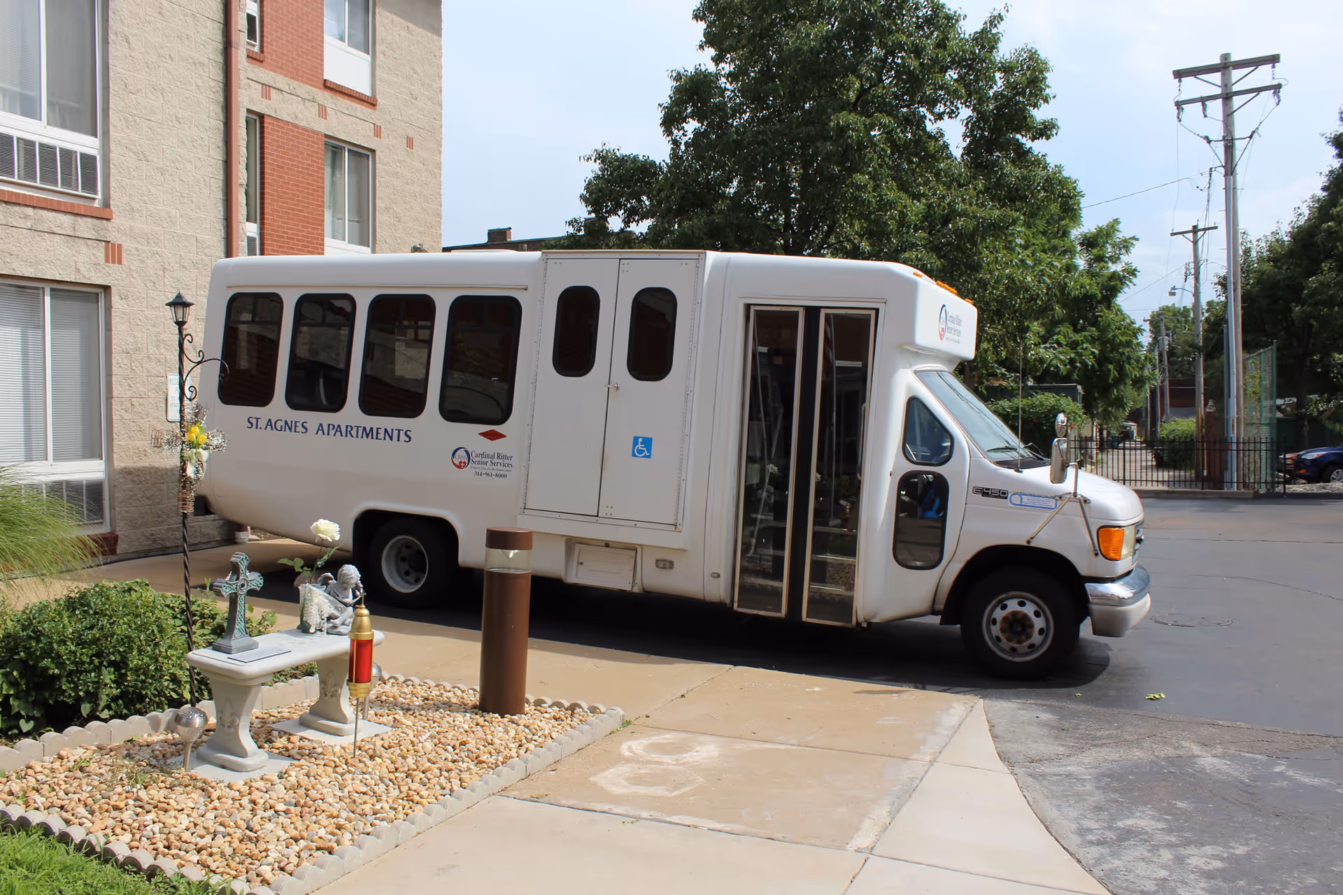 A white shuttle bus parked outside a building with the text 'St. Agnes Apartments' on its side. The bus has a handicap accessibility symbol near the door. In front of the bus, there is a small landscaped area with a bench, a cross, and some decorative items. Trees and power lines are visible in the background.