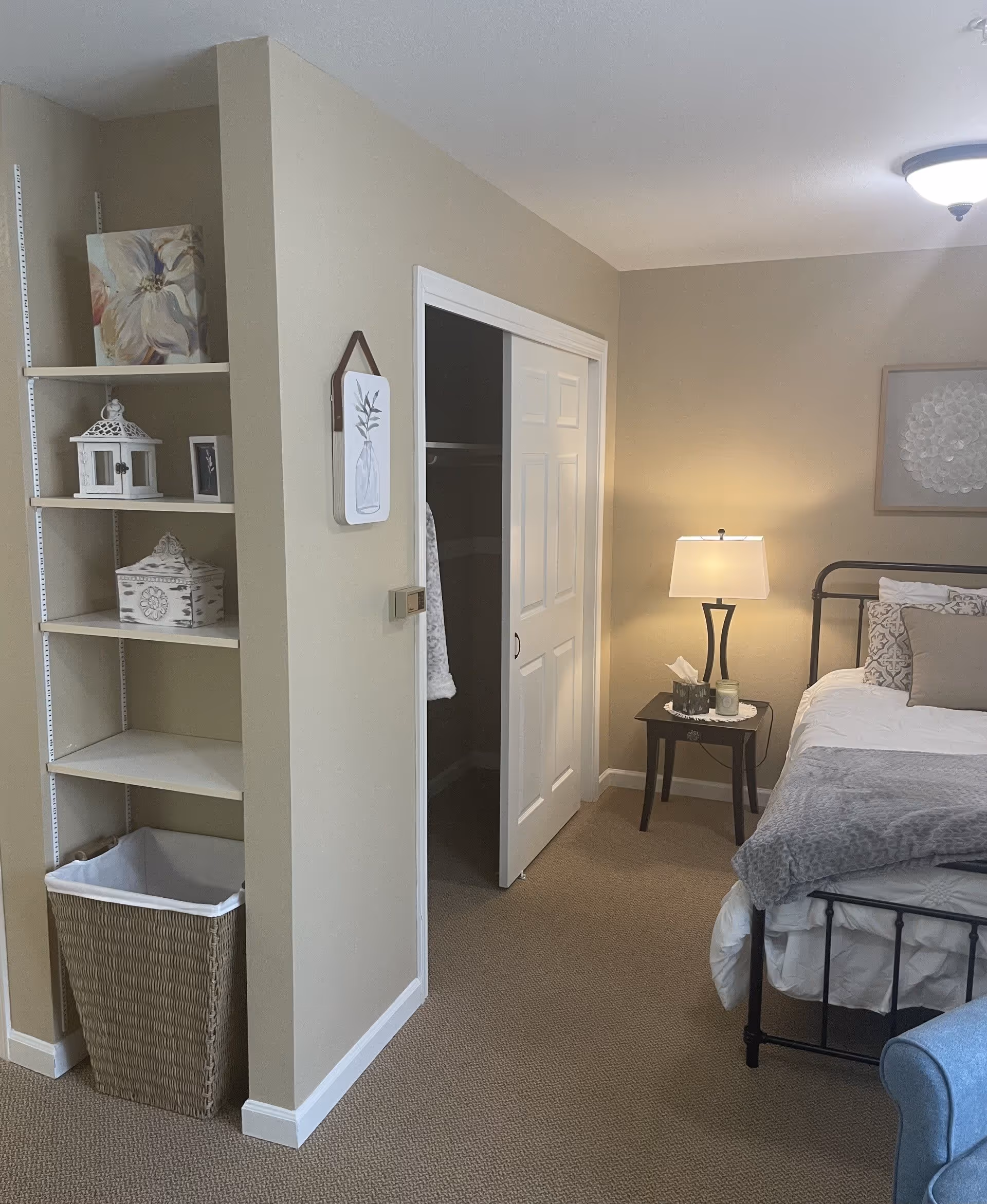 A cozy bedroom with beige walls and carpet. There is a bed with white and gray bedding and decorative pillows, a black metal bed frame, and a wooden nightstand with a lamp, tissue box, and candle. To the left, there is an open closet with white sliding doors and a shelving unit with decorative items and a wicker laundry basket below.