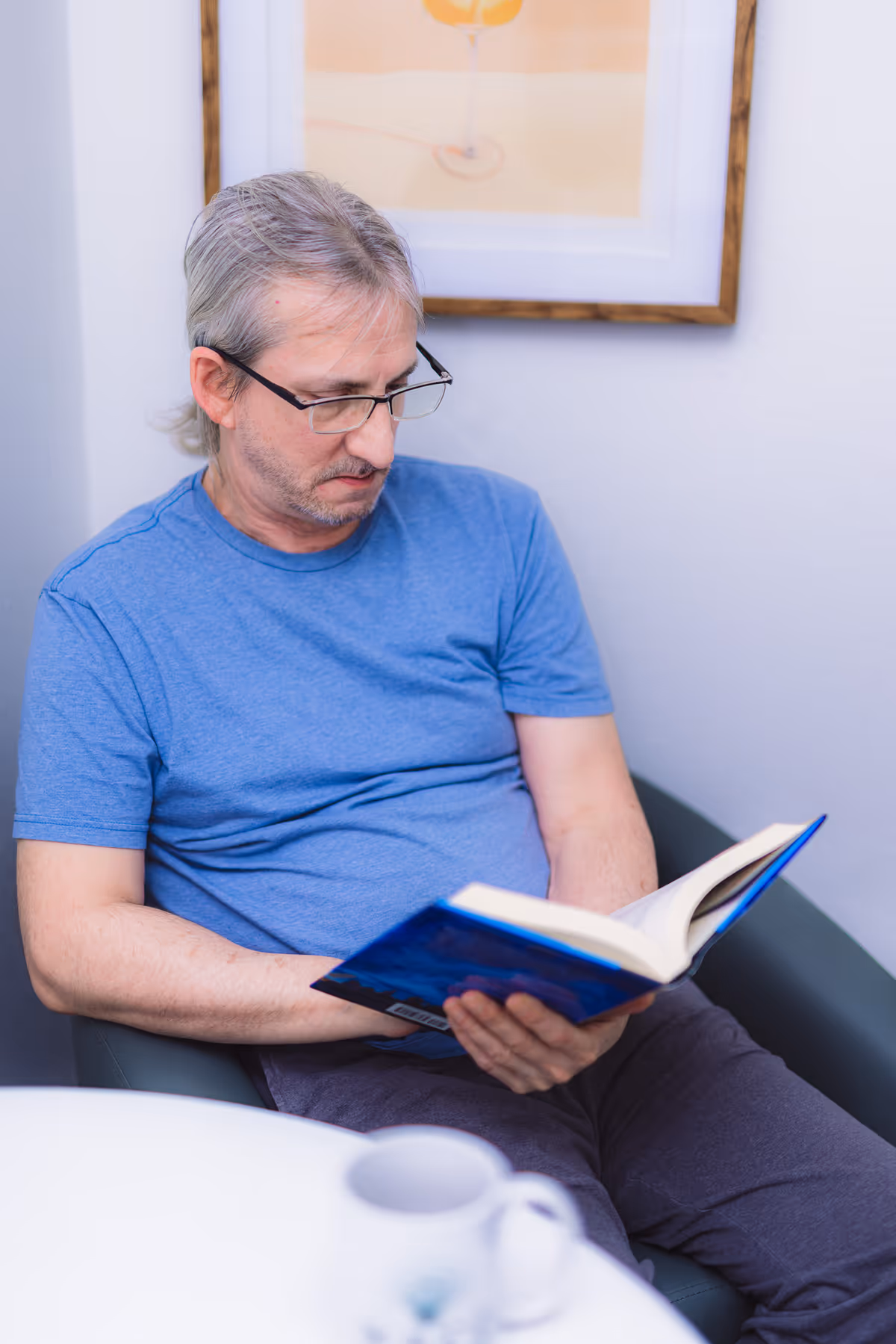 A middle-aged man with gray hair and glasses is sitting in a chair reading a book. He is wearing a blue t-shirt and dark pants. There is a white table with a white mug in front of him, and a framed picture of a drink on the wall behind him.
