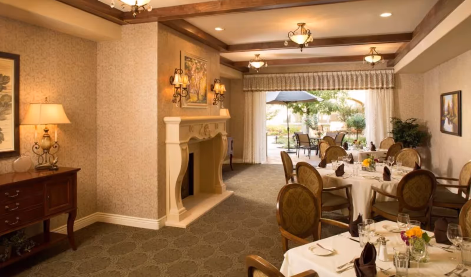 Elegant dining room with round tables set for a meal, a decorative fireplace, and an open view to an outdoor patio.