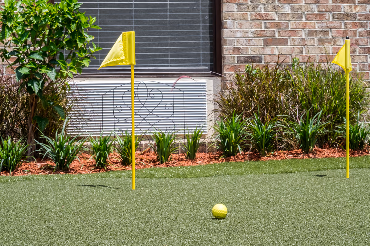 A small outdoor putting green with two yellow flags and a yellow golf ball on artificial turf, bordered by plants and a brick wall with a window and air conditioning unit.
