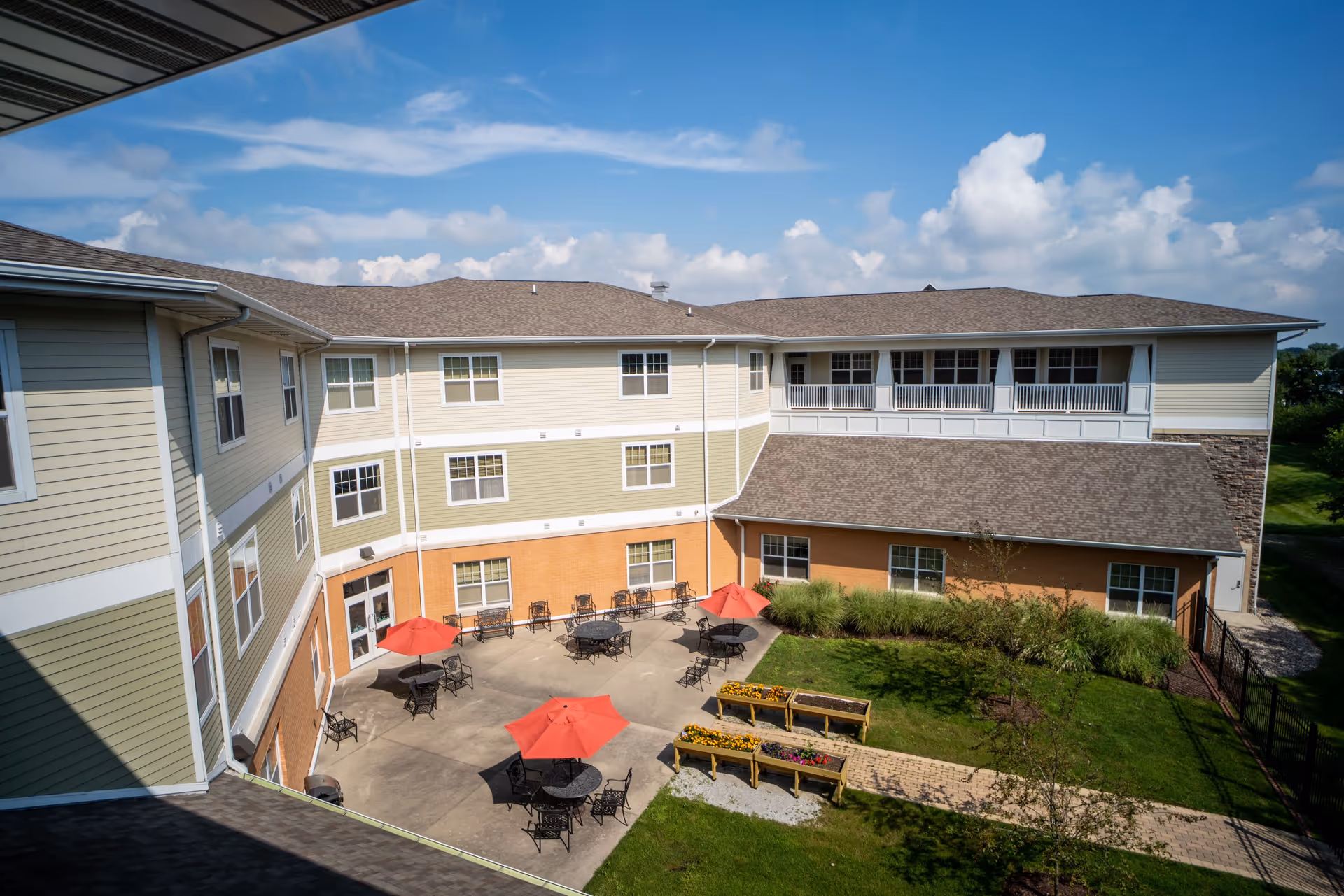 Outdoor courtyard area of a senior living facility with multiple round tables and chairs, some shaded by red umbrellas. The courtyard is surrounded by a three-story building with beige and light green siding and multiple windows. There are flower beds and a paved walkway in the grassy area adjacent to the courtyard under a partly cloudy sky.
