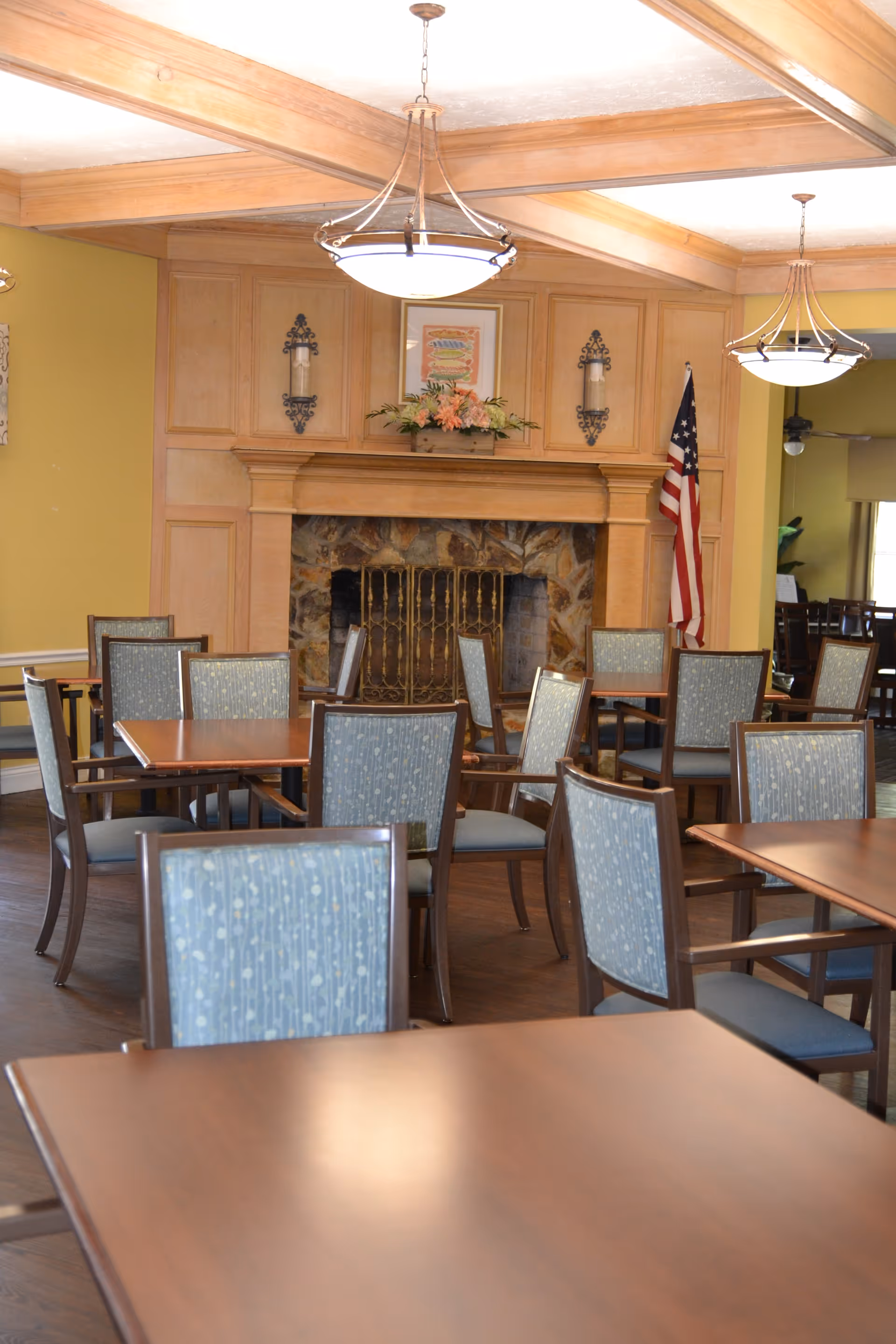 Dining area with multiple wooden tables and chairs featuring blue patterned upholstery. The room has a stone fireplace with a wooden mantel decorated with flowers and a framed picture above it. Two hanging light fixtures illuminate the space, and an American flag stands in the corner.