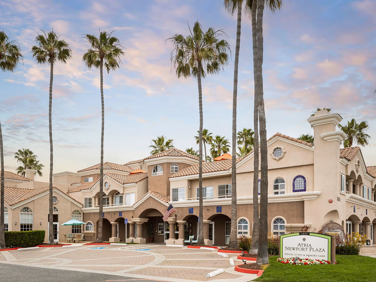 Exterior view of Atria Newport Plaza, a senior living and memory care facility, featuring a large building with beige stucco walls, red tile roof, tall palm trees, and a sign in front. The sky is partly cloudy with a soft sunset glow.