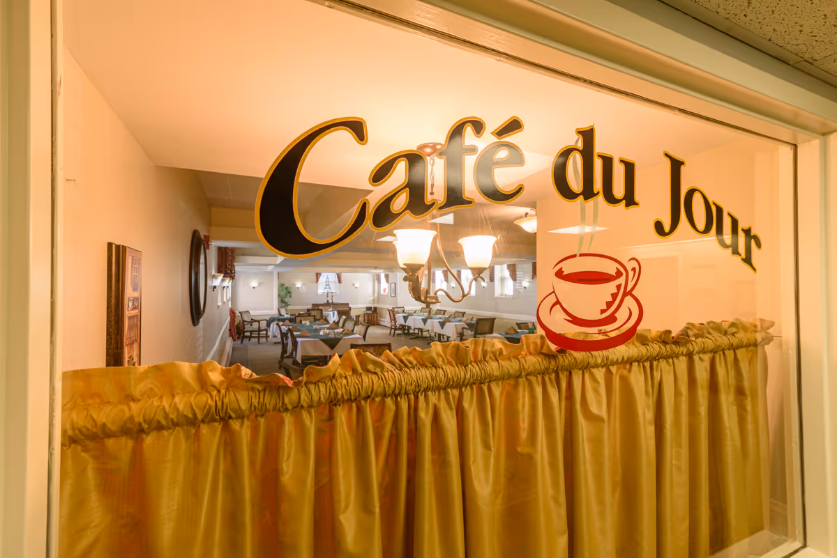 View through a window with the words 'Café du Jour' and a red coffee cup graphic, looking into a dining area with tables and chairs set up, illuminated by ceiling lights.