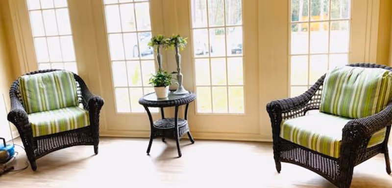 Two wicker armchairs with green and white striped cushions positioned on either side of a small round wicker table holding two potted plants, set in front of large glass-paneled doors letting in natural light.