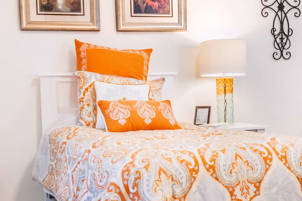 A neatly made bed with white and orange patterned bedding and multiple pillows in a bedroom. There is a white nightstand with a decorative lamp and a small framed picture next to the bed. Two framed artworks hang on the wall above the bed, and a decorative wrought iron wall piece is mounted on the adjacent wall.