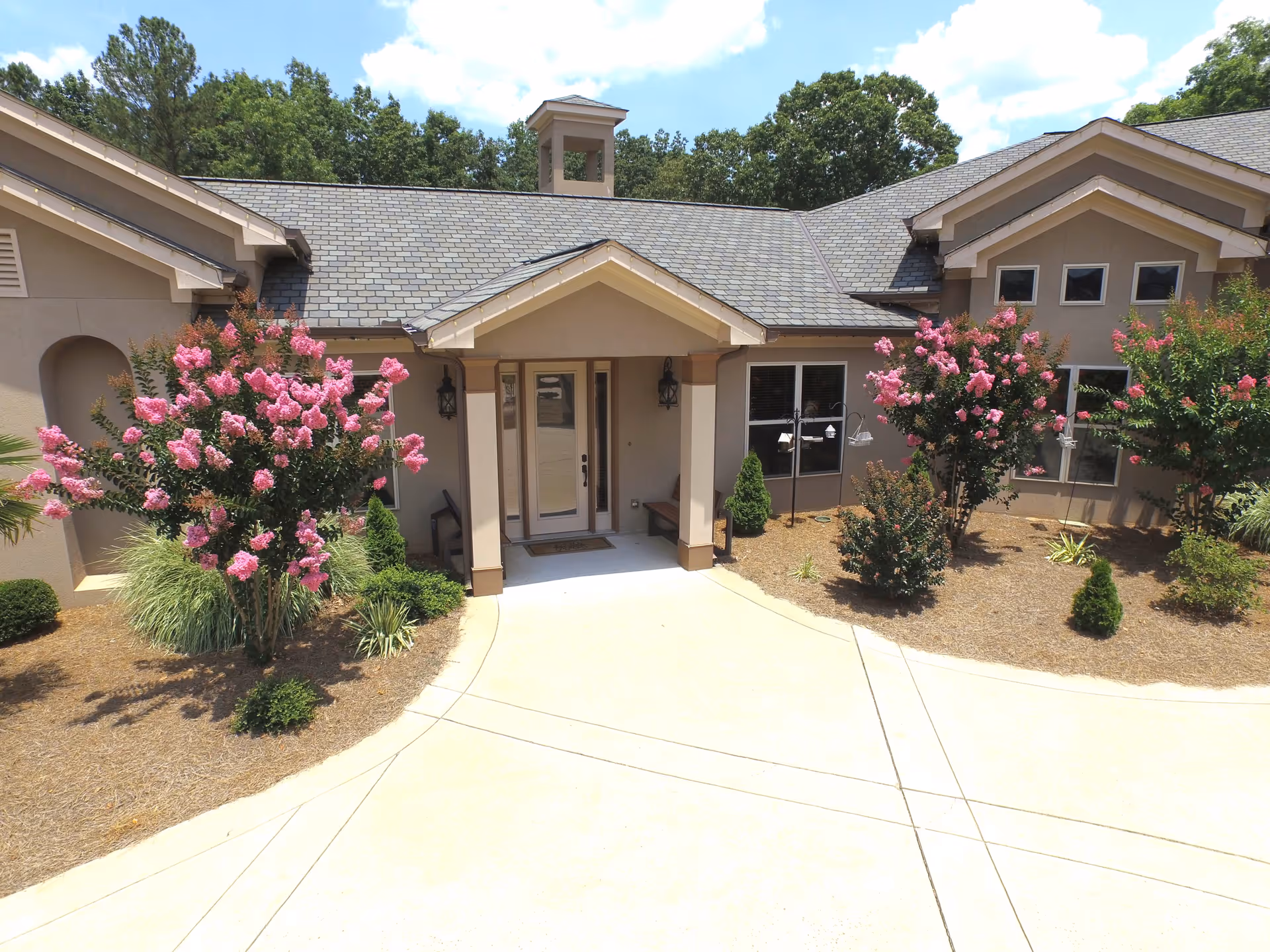 Front entrance of a senior living facility with a covered porch, beige exterior walls, and a gray shingled roof. The entrance is flanked by pink flowering bushes and landscaped with small shrubs and mulch. Trees and a clear blue sky are visible in the background.