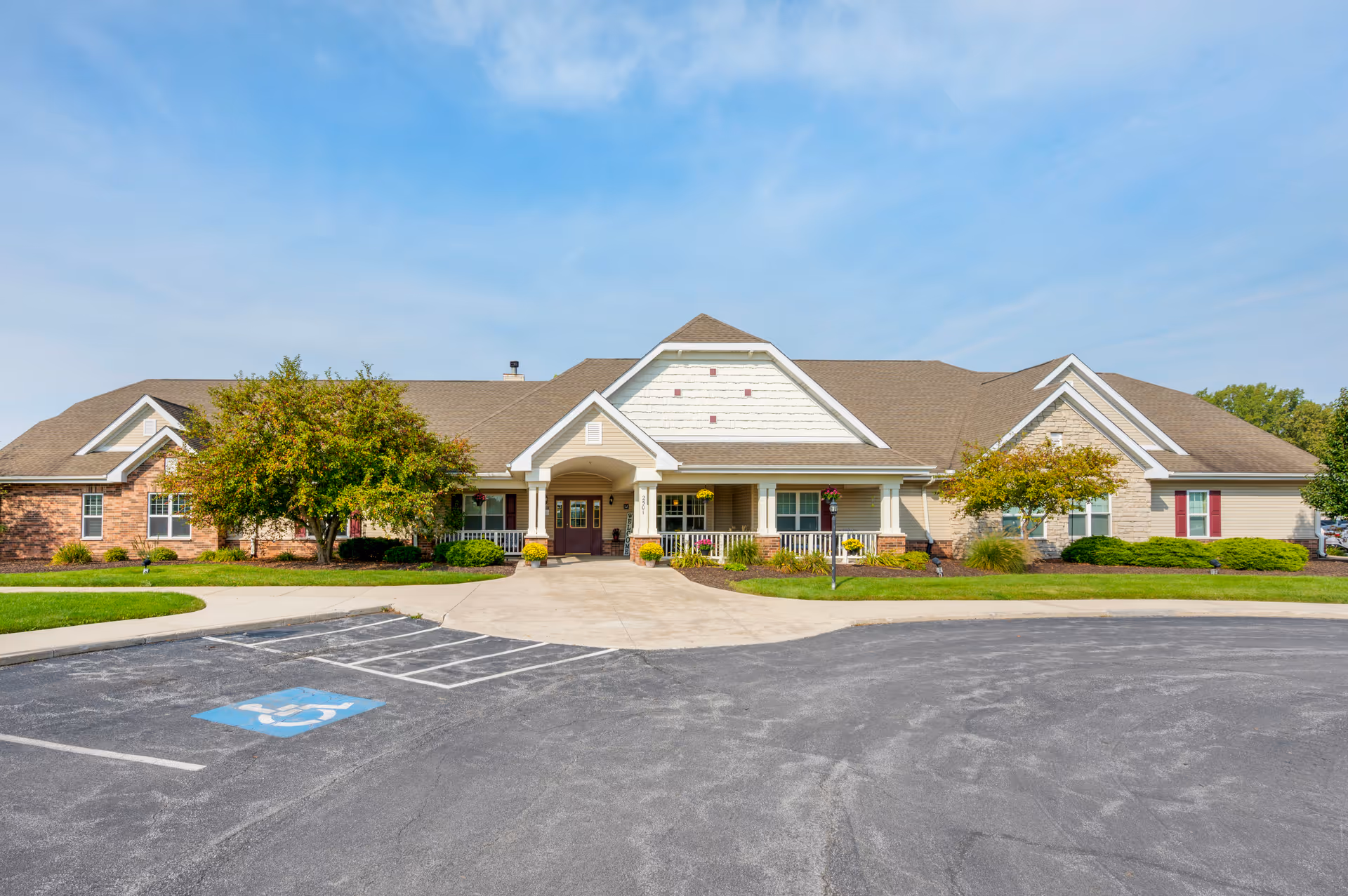 Front exterior view of a single-story senior living facility building with a covered entrance, surrounded by landscaped greenery and trees, under a clear blue sky. There is a parking lot with a handicapped parking space in front.