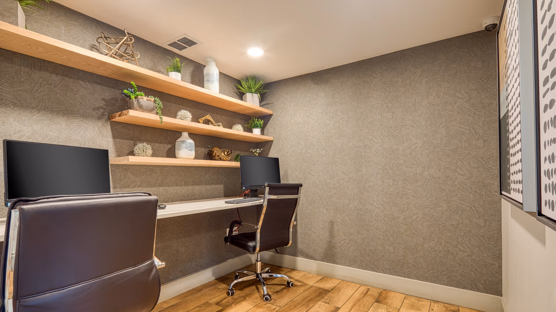 A small office or workspace with two black office chairs and two computer monitors on a white desk mounted to the wall. Above the desk are three wooden floating shelves decorated with plants, vases, and decorative objects. The walls have a textured beige wallpaper, and the floor is wooden. Two framed abstract artworks are partially visible on the right wall.