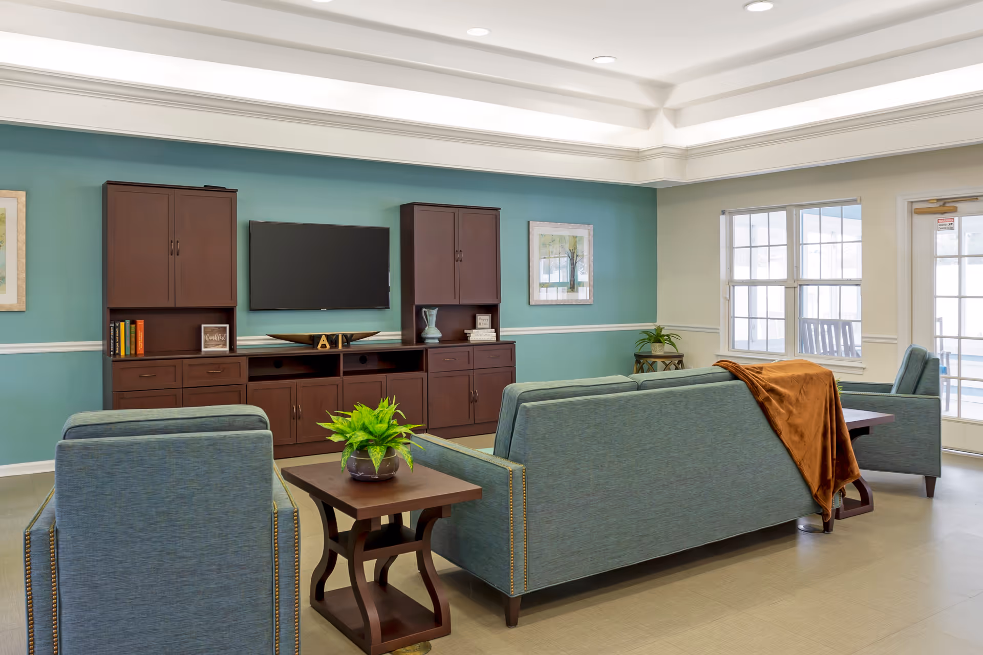 A cozy living room with teal upholstered sofas and armchairs arranged around a wooden coffee table with a green plant. The room features a teal accent wall with a mounted flat-screen TV and dark wooden cabinets on either side. There are framed pictures on the walls, large windows letting in natural light, and a door leading outside. A brown throw blanket is draped over the back of one sofa.