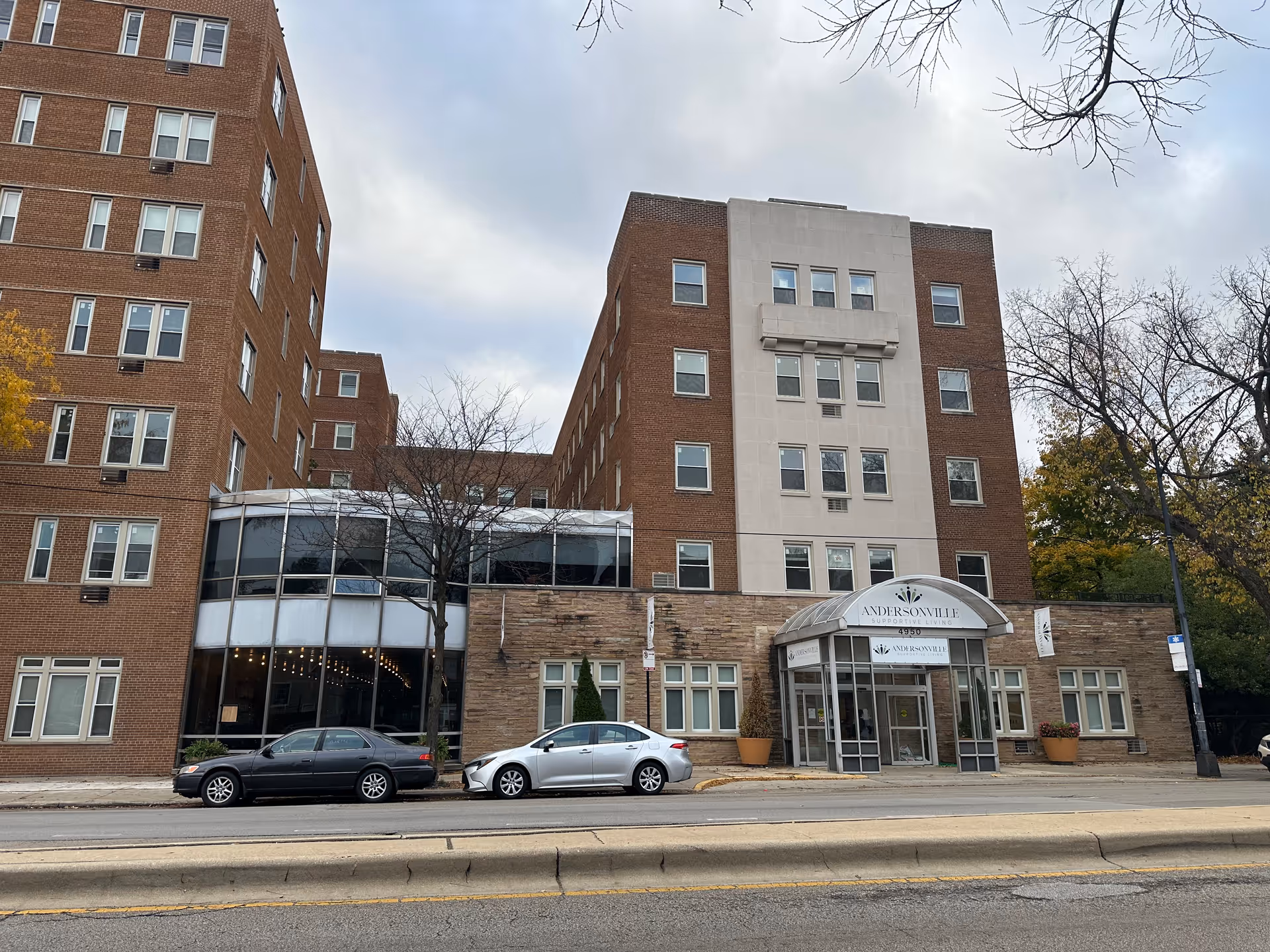 Exterior view of a multi-story brick building with a central entrance canopy labeled Andersonville Supportive Living. Two cars are parked on the street in front of the building, and leafless trees are visible around the area under a cloudy sky.