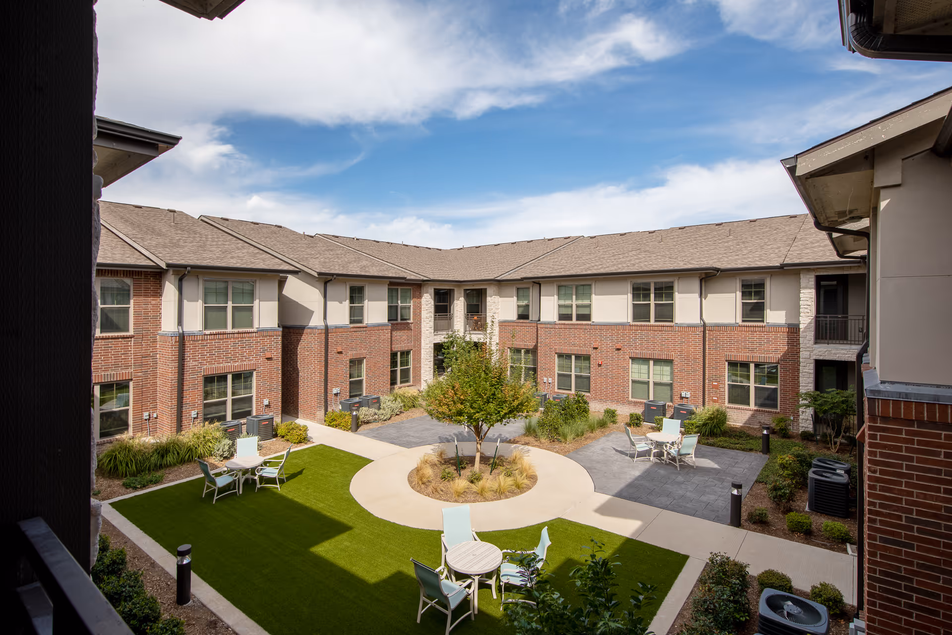 Landscaped central courtyard with seating, artificial turf, and a two-story brick senior living building under a blue sky.