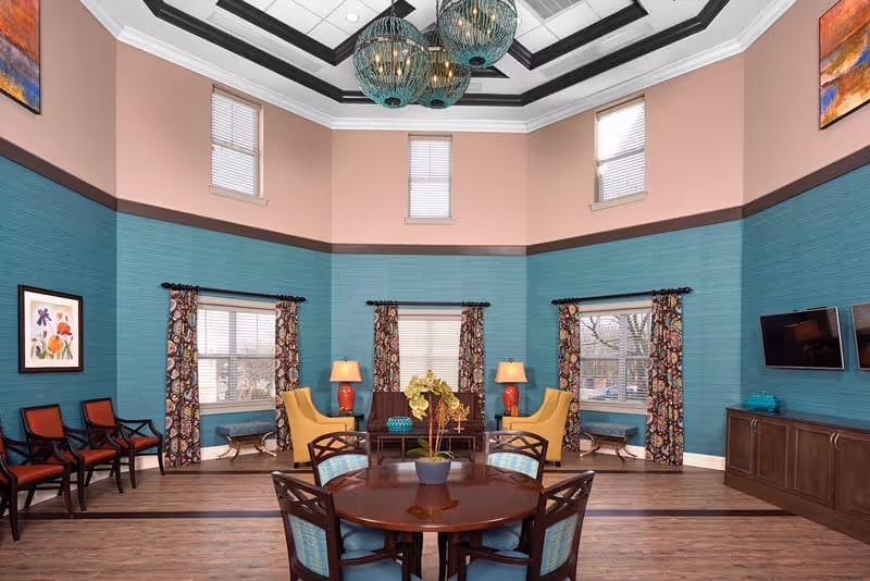 Spacious communal lounge with a round dining table, upholstered chairs and seating area against teal-accented walls beneath decorative pendant lights.