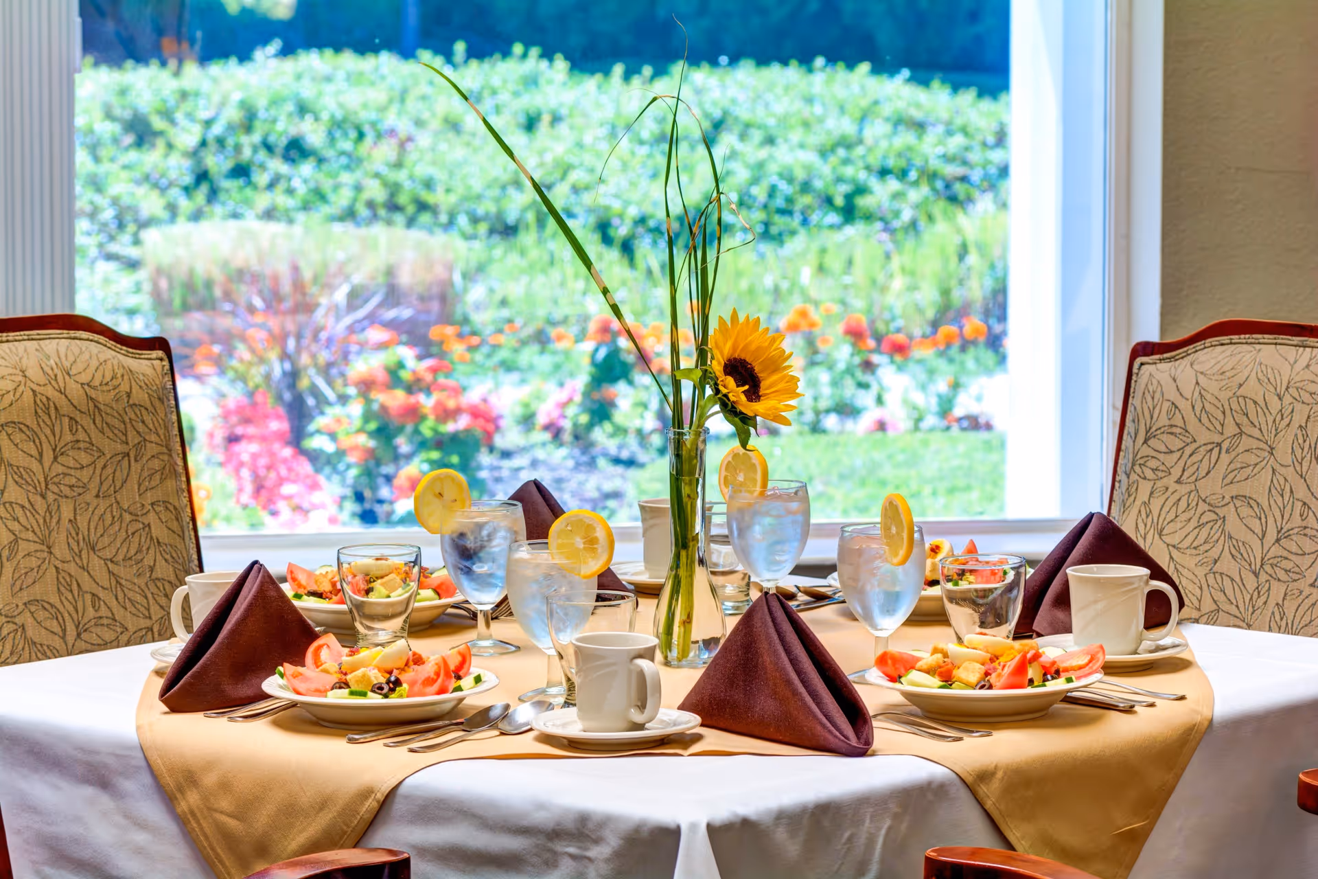 A dining table set for four with plates of fresh salad, glasses of water with lemon slices, coffee cups, and folded brown napkins. A vase with a sunflower and tall grass is in the center. The table is near a large window showing a colorful garden outside.