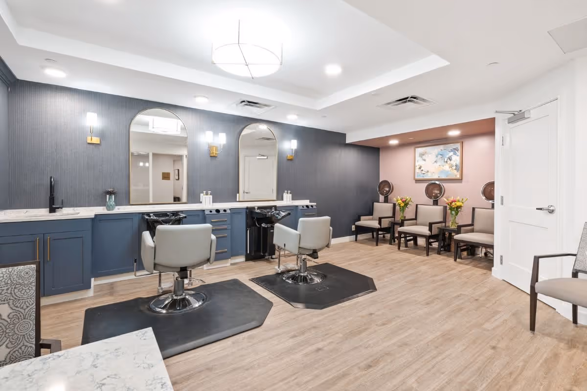 Interior view of a hair salon area in a senior living facility with two salon chairs in front of mirrors and sinks, a waiting area with chairs and hair dryers, light wood flooring, and modern lighting fixtures.