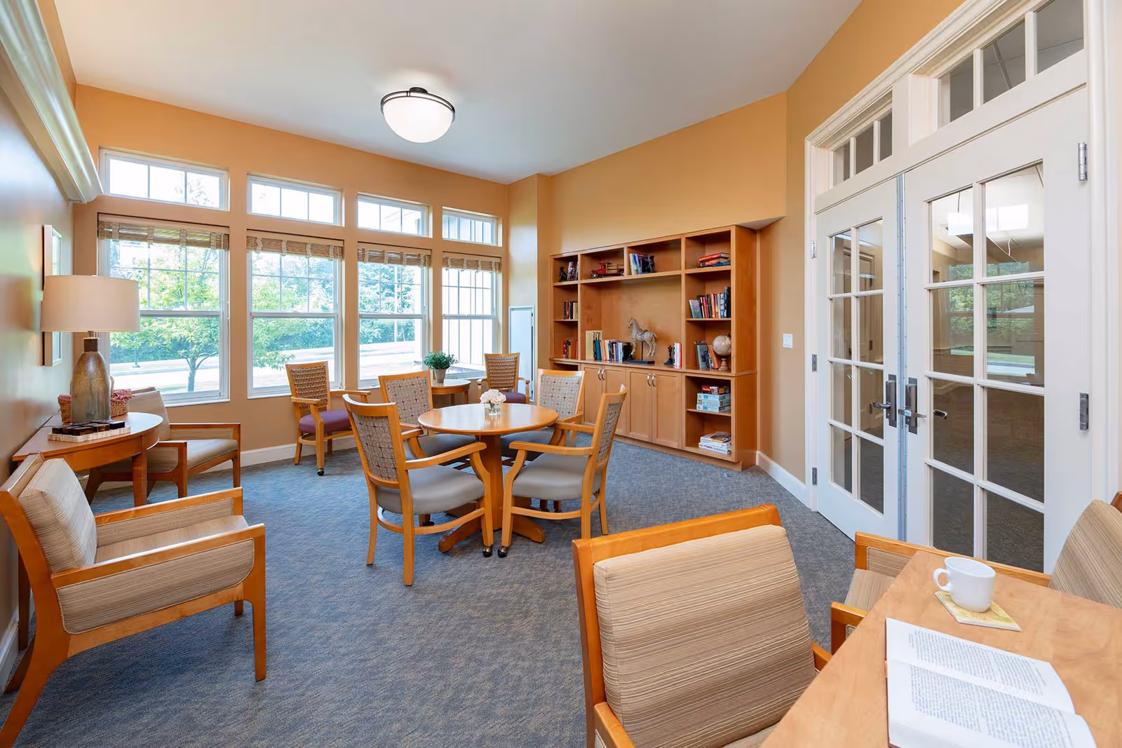 A bright and cozy common area with large windows letting in natural light. The room features several wooden chairs with cushions arranged around small round tables. There is a built-in wooden bookshelf filled with books and decorative items against one wall. A table with a cup and an open book is visible in the foreground. The walls are painted a warm beige color, and there are double glass doors leading to another room.