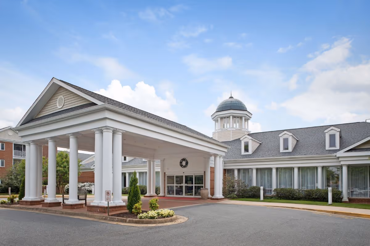 Front exterior view of The Colonnades facility featuring a covered entrance with white columns, a circular driveway, and a building with multiple windows and a cupola on the roof under a partly cloudy sky.