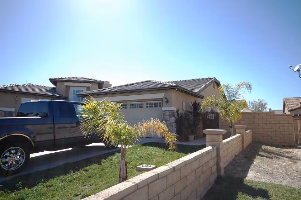 Exterior view of a single-story house with a tiled roof, a garage door, a black pickup truck parked in the driveway, a small front yard with green grass and palm trees, and a low brick wall along the front.