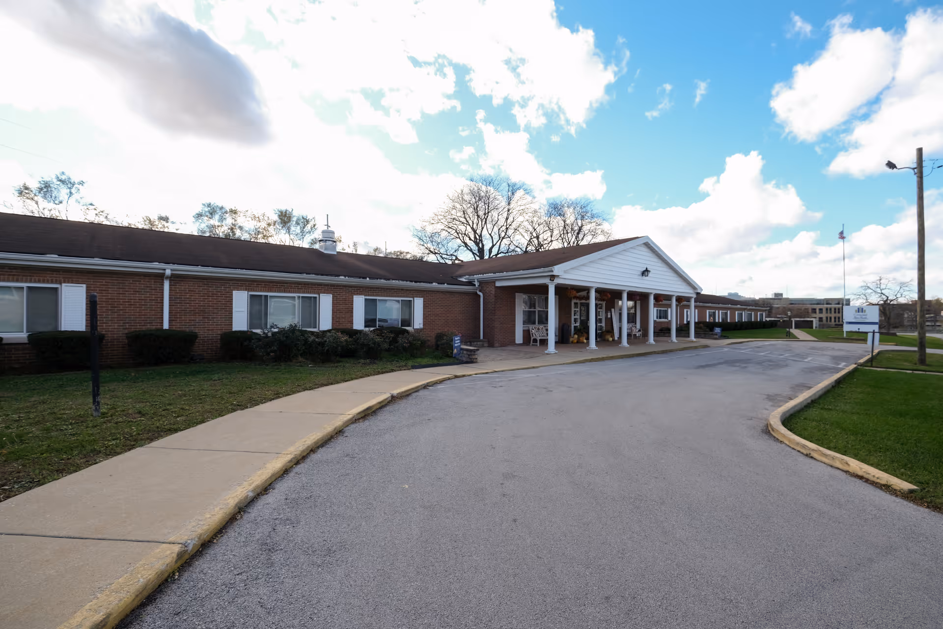 Single-story brick senior living facility with a covered entrance and circular driveway under a partly cloudy sky.
