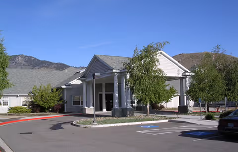 Front exterior view of a single-story building with a covered entrance supported by white columns, surrounded by trees and landscaping, with mountains visible in the background under a clear blue sky.