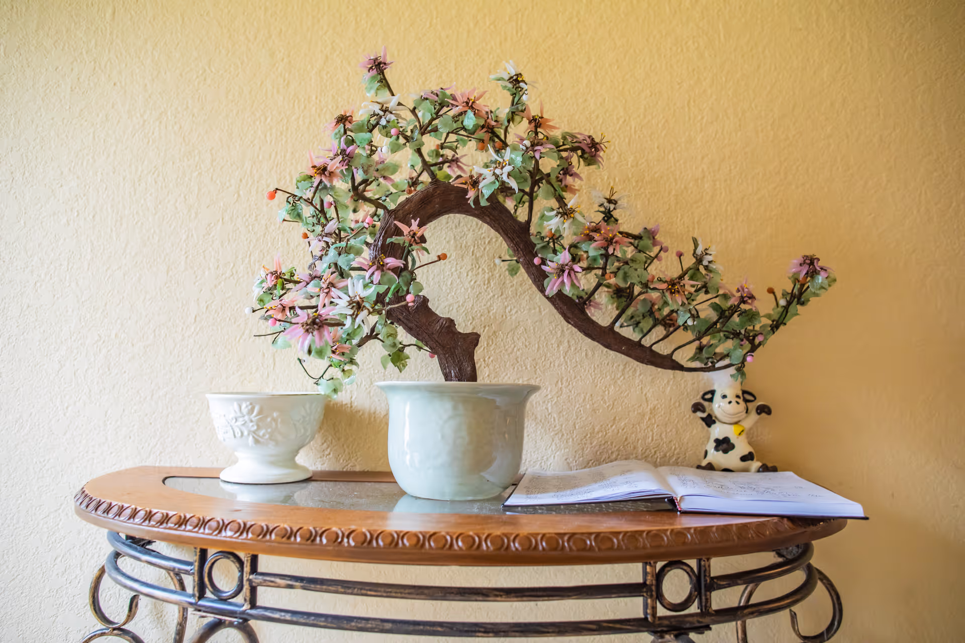 A decorative table against a beige wall with a potted artificial bonsai tree with pink and green flowers, a white ceramic bowl, an open book, and a small cow figurine.