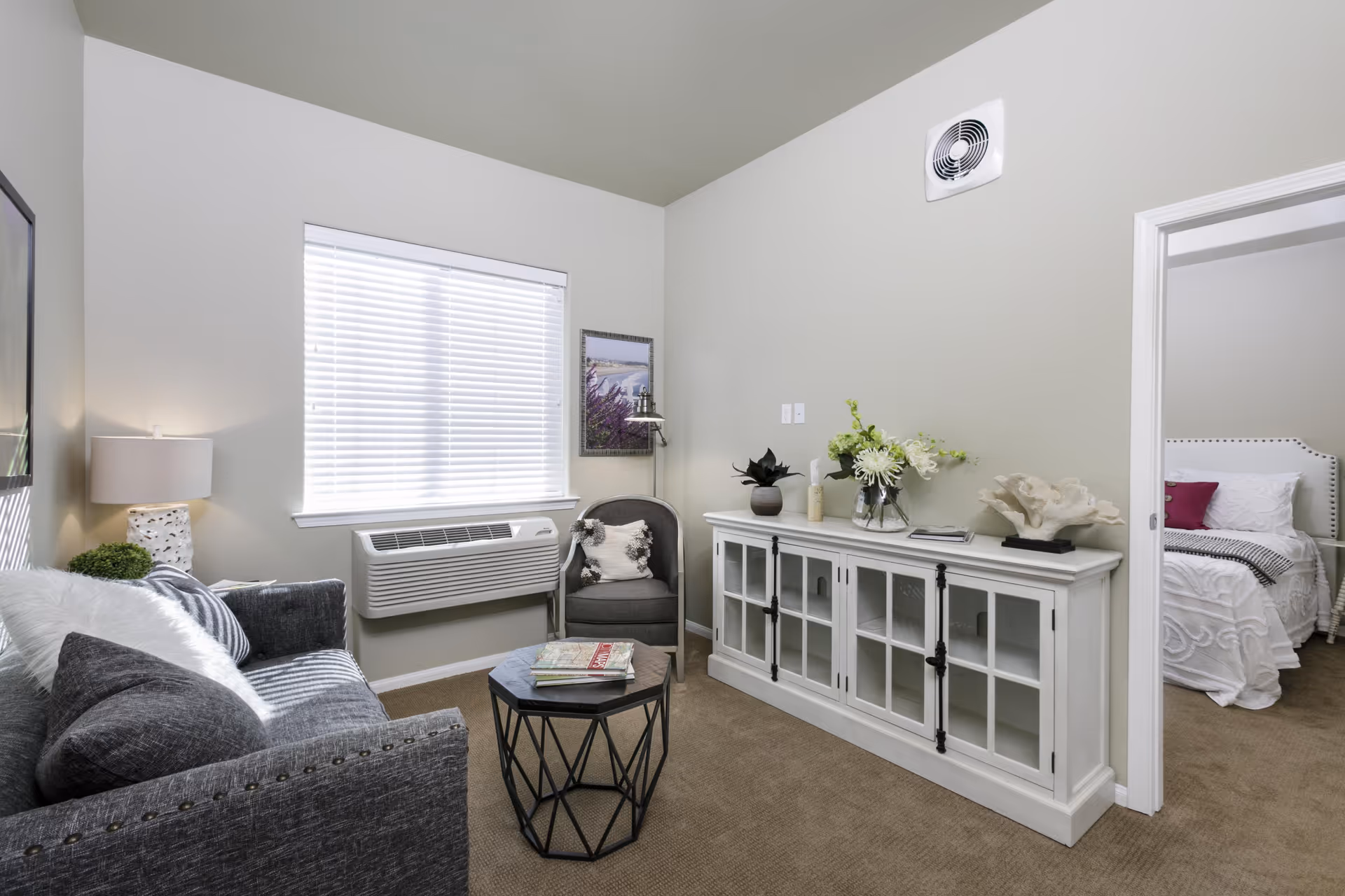 A cozy living room with a gray sofa adorned with pillows, a small round black coffee table with magazines, a gray armchair with a decorative pillow, a white cabinet with glass doors topped with plants and decorative items, a window with white blinds, and a glimpse into a bedroom with a white bed and pink pillow.