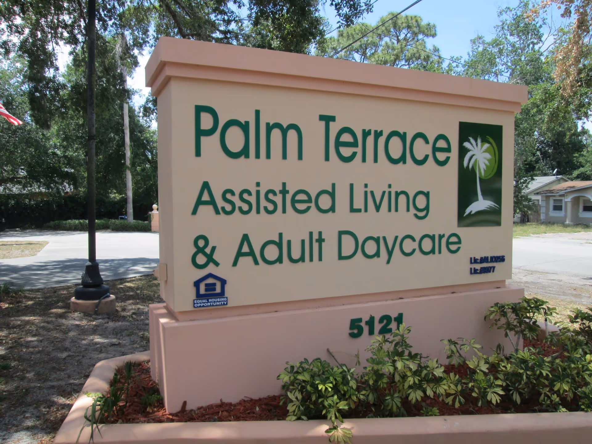 Outdoor sign for Palm Terrace Assisted Living & Adult Daycare Center with green text and a palm tree logo, surrounded by plants and trees in the background.