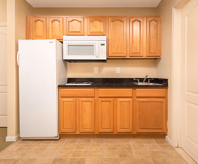 A small kitchen area with wooden cabinets, a white refrigerator on the left, a white microwave mounted above a black countertop, and a stainless steel sink on the right. The floor is tiled with light brown tiles and the walls are painted beige.